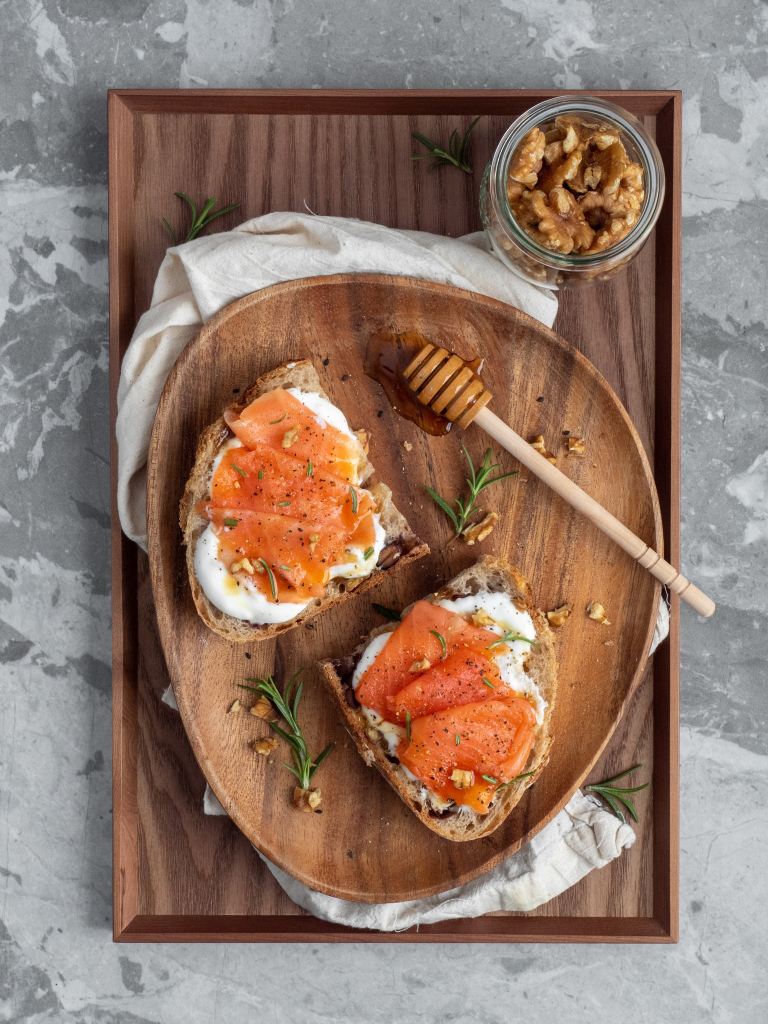 A wooden bowl, sat on top of muslin on top of a wooden tray. On the plate are two slices of bread witch cream cheese and smoked salmon on top. A jar of walnuts sit beside the plate.