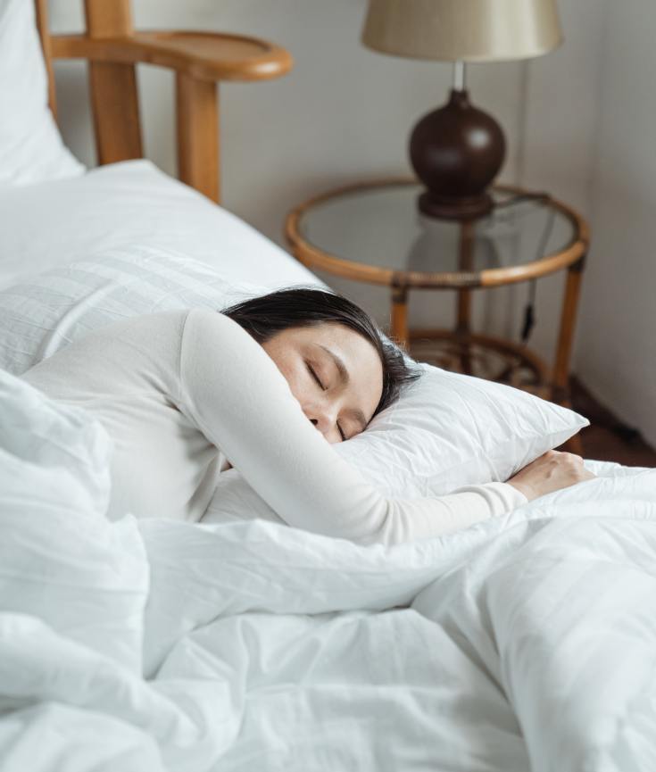 An Asian woman with dark hair wearing a long sleeve white top is sleeping on her side. The bedding is white and the bed frame is a medium tan wood. A glass and wicker side table is in the corner of the room with a beige lamp sitting on it. Blog post from Pandora's Health.