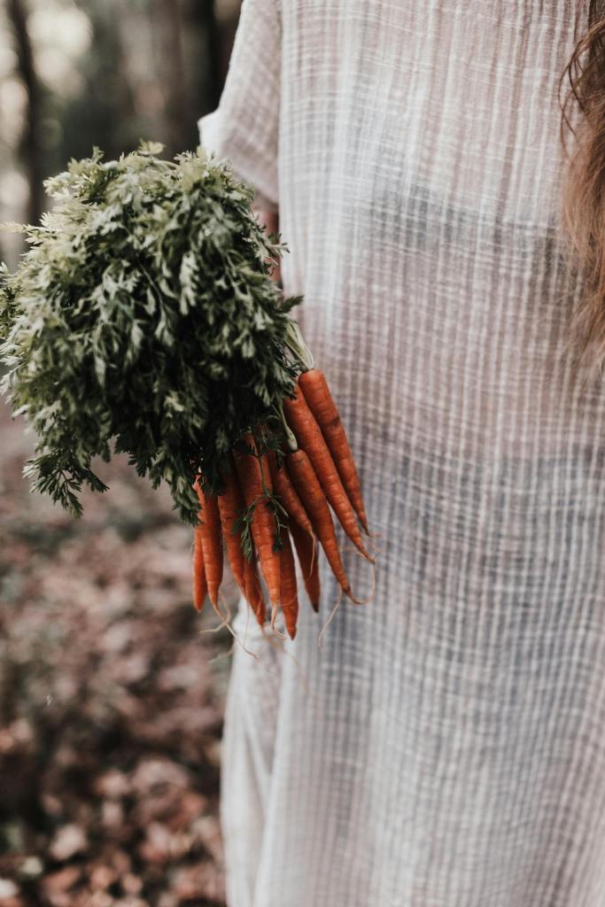 A woman in a linen dress is holding a bunch of carrots, which include their green tops. Pandora's Health blog.